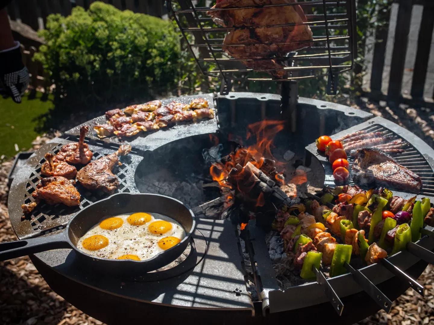 Remundi Feuerstelle mit Grillmodulen rund um das Feuer: Pfanne mit Spiegeleiern, Spieße, Grillrost und Plattensegmente