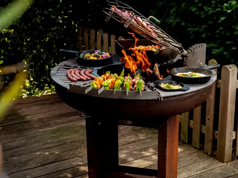 Remundi Carus L Modulgrill auf einer Holzterrasse, bestückt mit Pfannen, Spießhalter und Grillplatten-Modulen rund um das Feuer